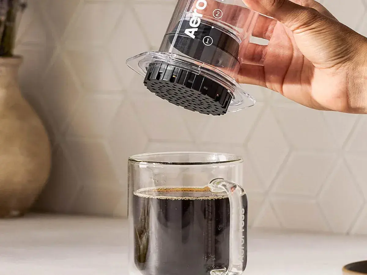 Hand lifting a clear AeroPress off a glass mug of coffee